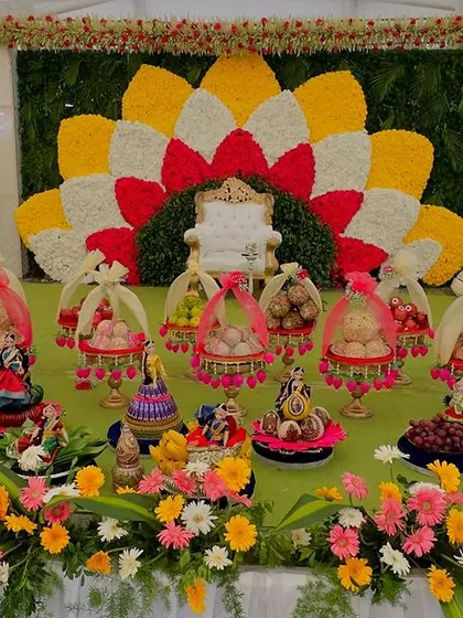A wide view of a platter decoration setup for a half-saree function. The arrangement includes gift baskets, doll platters, and fruit trays, all beautifully laid out in front of a floral backdrop.