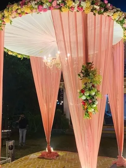 A beautiful and romantic outdoor wedding mandap. The design features a circular structure with delicate peach-colored drapes, a classic chandelier for evening ceremonies, and lush floral arrangements, creating a perfect setting for your vows.