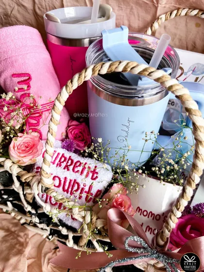 A top-down view of the baby shower basket, showing the personalized 'Mama's Daddy' sipper, embroidered towel, and fresh flowers.