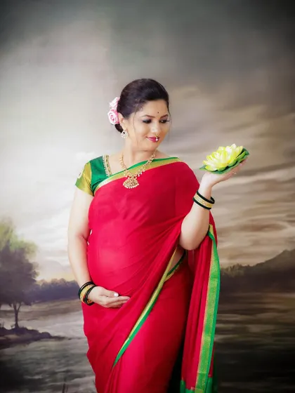 A classic portrait celebrating tradition. The mom-to-be is dressed in a beautiful red and green saree, holding a lotus flower against a painted backdrop.