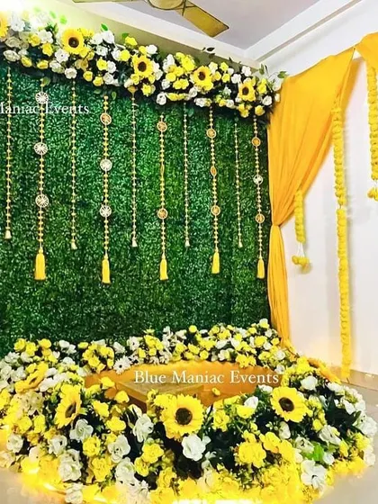 A Haldi and Mehendi setup featuring a green wall with hanging tassels and a base of sunflowers. The yellow drapes and marigold garlands add a traditional touch.