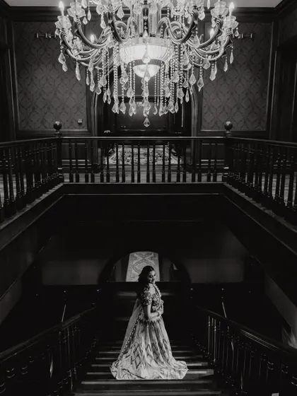 Another artistic black and white shot from the Sangeet. This full-length portrait captures the bride's elegance against the dark, moody backdrop of the grand staircase, creating a feeling of timeless romance.