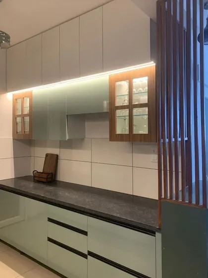 A serene kitchen design with light green cabinets and a dark countertop. The wooden-framed glass cabinets and the slatted wood partition add warmth and texture to the space.