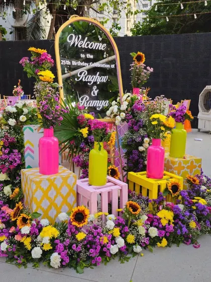 A colorful welcome sign for a twins' 25th birthday celebration, surrounded by a vibrant mix of sunflowers and purple and pink flowers.