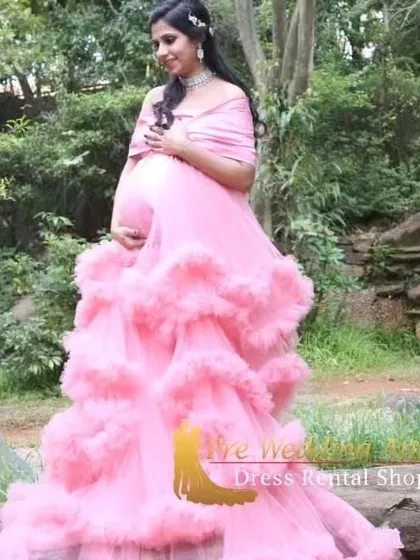 The soft pink ruffled gown looks ethereal in a lush, green setting. The off-the-shoulder design and layers of tulle create a romantic and timeless look for any expecting mother.