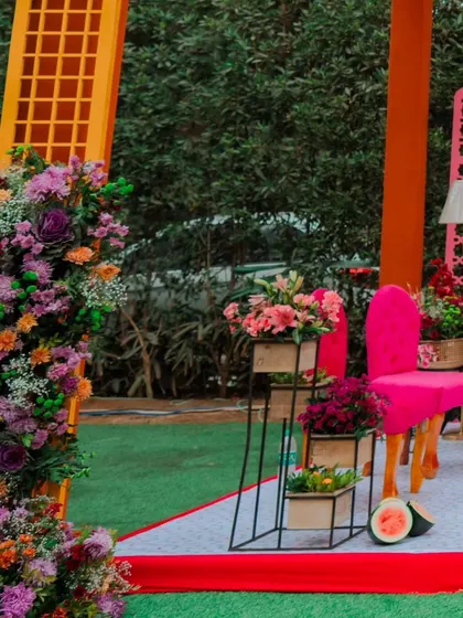 A side view of the Mehendi stage, showcasing the layers of colour from the orange structure, pink jaali, and lush, multi-coloured floral arrangements.