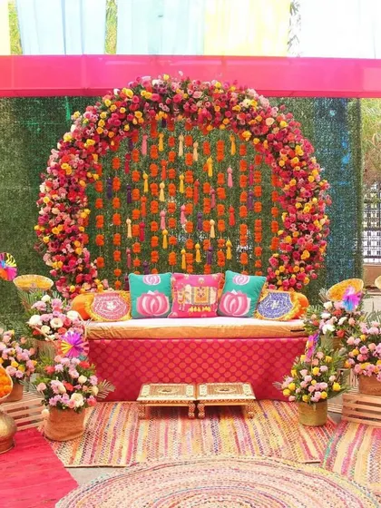 A vibrant and colorful Mehendi stage. The circular floral arch is filled with bright flowers, and the seating is adorned with colorful cushions, creating a joyful atmosphere.
