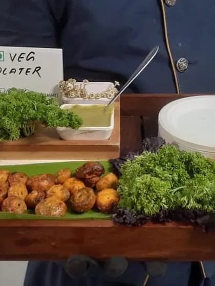 A server presents a vegetarian platter of roasted baby potatoes. The dish is served on a wooden tray with fresh parsley and a side of green chutney, lit by a candle for a warm and inviting touch.