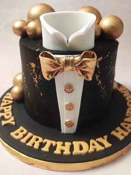 We present the essence of formal sophistication with our black and gold tuxedo cake. This design features a sharp, edible shirt collar and a lustrous gold bow tie set against a matte black tier, accented with gold splatters and spheres, making it an impeccable choice for a gentleman's milestone birthday.