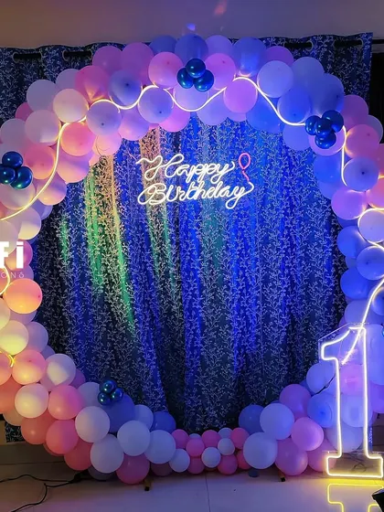 This first birthday photo booth features a circular balloon backdrop in pink and blue. The design is enhanced with a 'Happy Birthday' neon sign, a lit-up number '1', and a backdrop of artificial green leaves for a touch of texture.