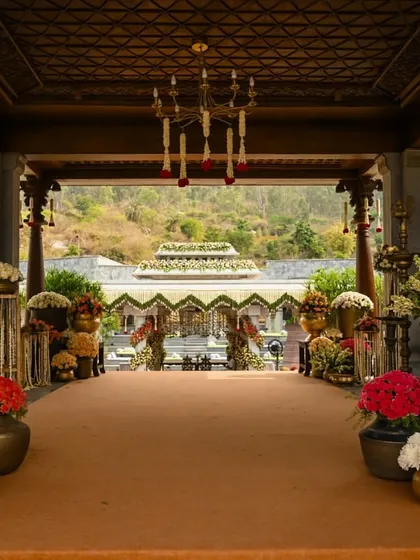The grand entrance to a wedding venue, lined with an abundance of colorful flowers in traditional brass pots, leading to a stunning mandap in the distance.
