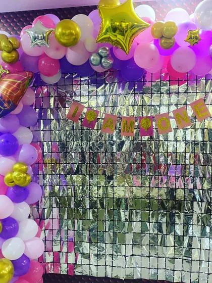 A home birthday decoration with a butterfly theme. The setup includes a silver foil curtain backdrop, a balloon arch, and a "I AM ONE" banner.