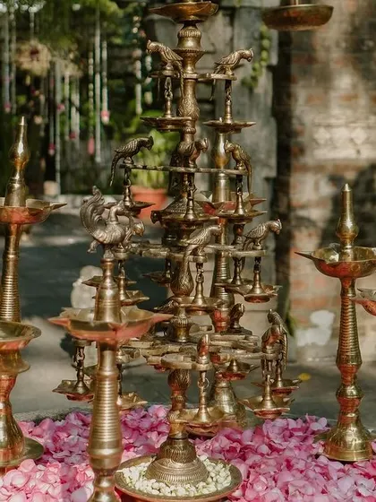 A collection of traditional brass lamps (diyas) of various sizes are arranged on a bed of pink rose petals, ready to be lit for the ceremony.