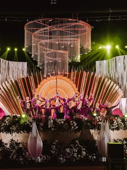 A wide shot of a grand sangeet stage we designed and choreographed. The stage features a massive, ornate sun dial backdrop, floral arrangements, and stunning chandeliers, with our dancers in formation, ready to begin a fairytale performance.