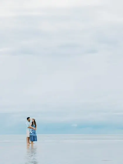 A wide-angle shot capturing the couple standing in the shallow waters, emphasizing the tranquility and beauty of the beach location.