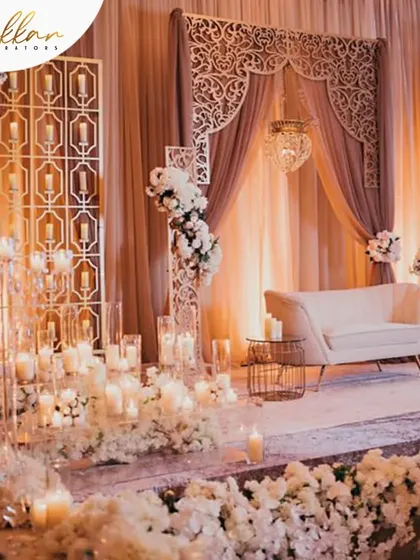 An all-white reception stage that exudes serenity and elegance. The decor features a wall of white flowers, candles, and intricate laser-cut panels for a modern yet timeless feel.