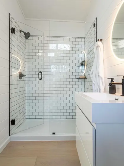 This bathroom design features a clean, modern aesthetic with white subway tiles and a glass shower enclosure. The illuminated mirror adds a touch of sophistication and provides excellent lighting.
