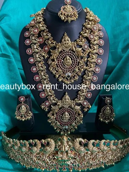 A grand bridal set with intricate peacock and Lakshmi motifs, studded with red stones. This rental comes with a choker, long necklace, vaddanam, and maang tikka.