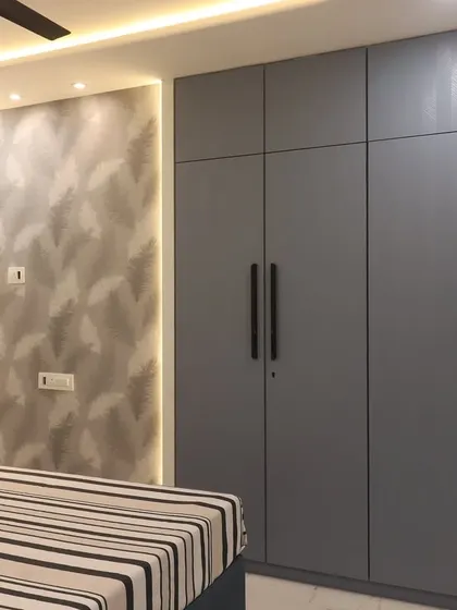 A master bedroom in Bhayandar West with a cool grey color palette. The room includes a large four-door wardrobe, a textured wallpaper accent wall with backlighting, and open shelves for displaying personal items.