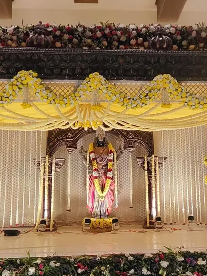 This is a traditional South Indian wedding mandap I built around a central deity statue. The design uses yellow and white fresh flowers for the garlands and pillar decorations, set against a classic carved wooden frame.
