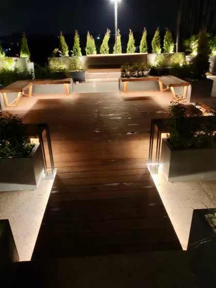 A rooftop garden at night. The wooden deck is flanked by benches and planters, with subtle under lighting creating a sophisticated and inviting pathway.