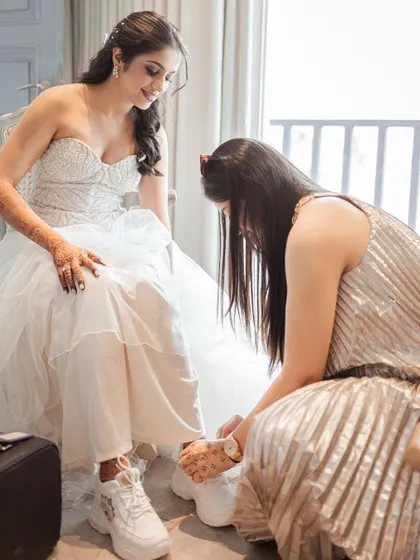 A bridesmaid helps the bride with her stylish white sneakers, a perfect blend of elegance and comfort. It's these small, supportive moments with the bride tribe that make the getting-ready process so special.