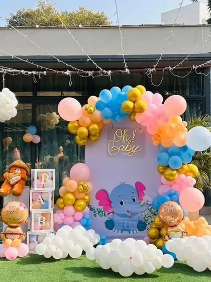 An outdoor baby shower with a cute elephant theme. This setup includes a custom 'Oh! Baby' backdrop, a colorful organic balloon arch in pastel and gold, and themed props like a teddy bear and letter blocks.