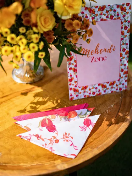 A 'Mehendi Zone' sign on a wooden table, with custom-printed napkins ready for guests.