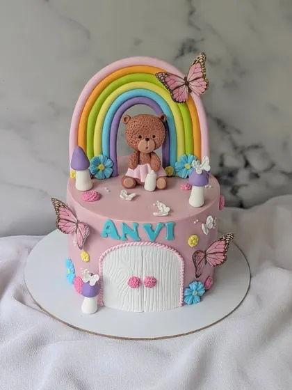 A sweet slice of sunshine for little Anvi. This cake features a teddy bear's fairy house, complete with a rainbow, butterflies, and toadstools.