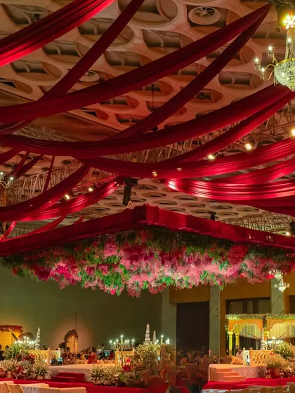 A wide view of a reception hall with a ceiling draped in red fabric and a massive floral chandelier at its center. The design creates a sense of grandeur and celebration.