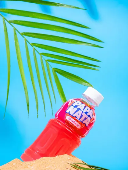 A tropical-themed shot of a Papa Nata strawberry drink, styled with sand and a palm leaf. This creates a vacation vibe, associating the beverage with refreshment and fun.