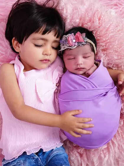 A heart-melting moment of pure love as an older sister sleeps beside her swaddled newborn sibling on a soft pink blanket.