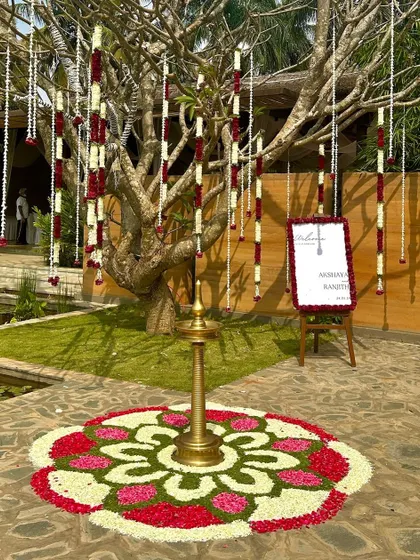 Another stunning floral rangoli in red, white, and green, with a tall brass lamp and hanging flower strings in the background.