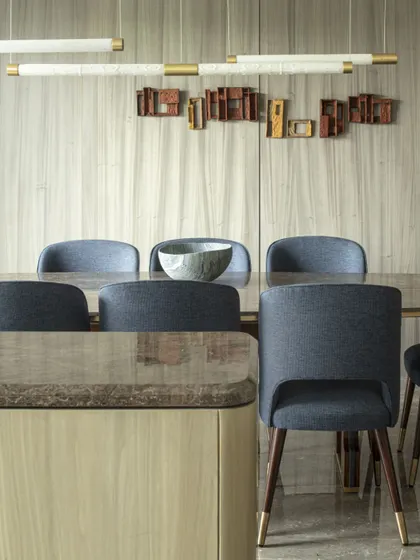 The dining area of the Jor Bagh Residence is highlighted by a dazzling brass installation and exquisite Makrana marble lights. Midnight blue upholstered chairs seamlessly offset the muted color palette of the wood-paneled walls.