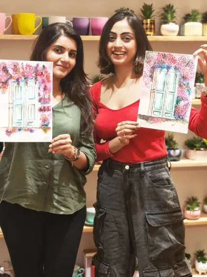 Two women happily hold up their matching watercolor paintings of a charming door framed by flowers, created in my Pune studio.