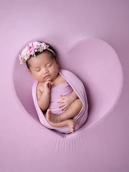 A creative variation of the swaddled pose, with the baby nestled inside a heart-shaped cutout on a purple backdrop.
