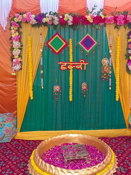 A traditional Haldi ceremony setup with a large brass urli filled with rose petals. The backdrop features green and yellow drapes, marigold garlands, and colorful hangings.