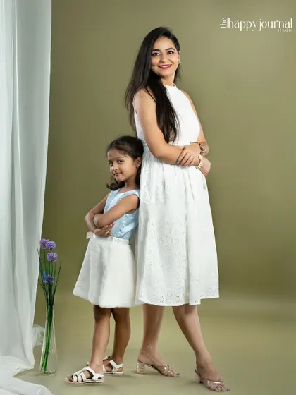 A mother and daughter stand back-to-back, striking a fun and confident pose. Their matching white dresses and playful attitude make for a memorable portrait.