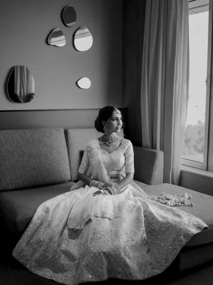 A classic black and white bridal portrait, with the bride seated elegantly on a sofa by a window.