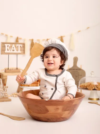 A happy chef in his kitchen. This little one holds up a wooden spoon with a big smile, surrounded by baked goods and kitchen props in this delightful themed shoot.