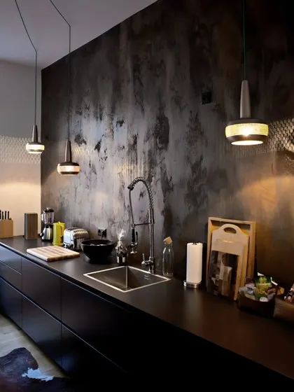 A moody and industrial-inspired kitchen with all-black cabinetry and a textured concrete-effect wall. The minimalist pendant lights provide focused task lighting over the sleek, dark countertop.