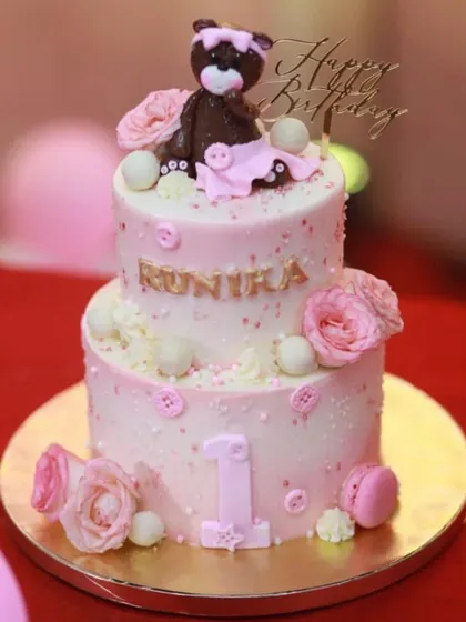 A lovely two-tiered pink cake for Runika's first birthday. This design features a fondant teddy bear, pink roses, macarons, and the number "1" prominently displayed on the bottom tier.