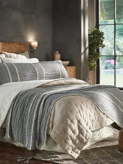 A cozy, rustic bedroom scene with a rattan headboard and layered neutral bedding. The natural light from the large window creates a bright and airy feel, highlighting the textures of the quilts and throws.