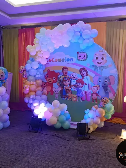 A Cocomelon-themed stage in a banquet hall. The large circular backdrop is framed by a pastel balloon arch and complemented by a light-up number "1" and character props.