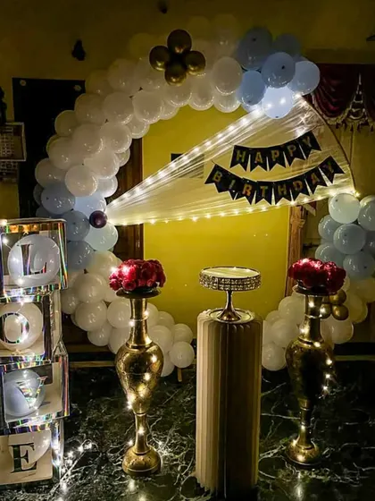 We create loving tributes for your special occasions. This home setup features a blue and white balloon arch, elegant brass vases with red roses, and illuminated "LOVE" display boxes for a heartfelt anniversary or birthday celebration.