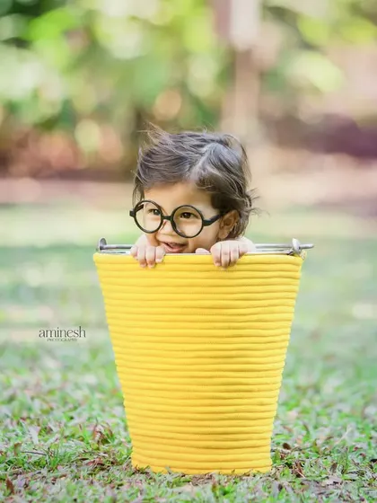 A playful outdoor shot of a curious baby with glasses peeking out of a bright yellow bucket. This type of fun and creative kids' portrait captures their charming personality.