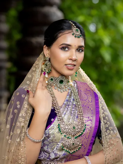 A classic Telugu bride look featuring a purple silk saree with a golden border and a sheer veil. The emerald jewelry adds a touch of regal elegance.
