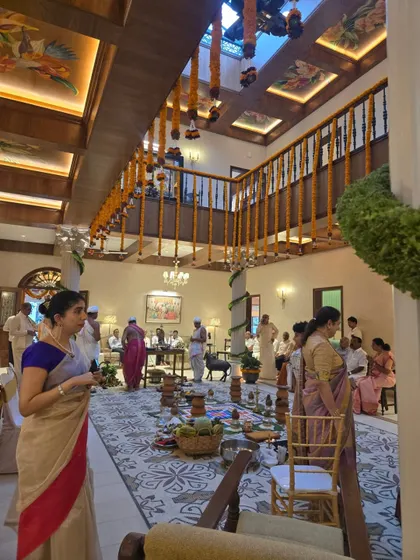 The spacious, open-plan living area of the Veerabhavana Villa during a housewarming ceremony. The design features a coffered ceiling with painted details, a large patterned rug, and a wooden balustrade, creating a home perfect for large family gatherings.