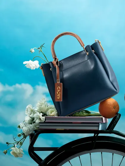 A whimsical and fresh shot of a navy blue handbag resting in a bicycle basket with flowers. This lifestyle image tells a story of a chic day out, making the product feel aspirational and stylish.