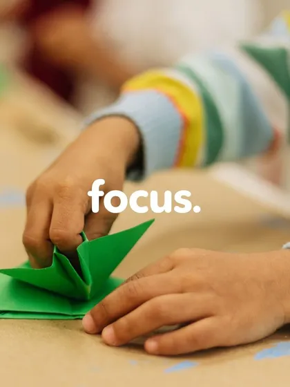 Focus in action. A child's hands are seen carefully folding a piece of green paper. This origami activity is a fantastic way to improve concentration, patience, and spatial reasoning skills.